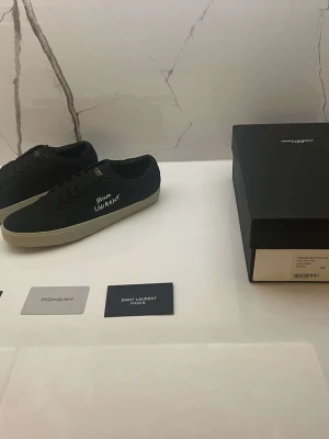 Svarta Saint Laurent sneakers - Snygga svarta sneakers från Saint Laurent med vit sula och logga på sidan. Klassisk låg modell med snörning och minimalistisk design. Perfekta för dig som gillar stilrena och exklusiva skor. Materialet är canvas och detaljerna är diskreta men lyxiga.