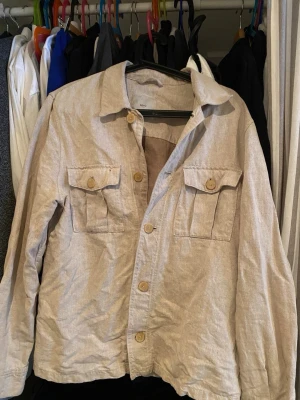 Beige overshirt från H&M - Säljer en beige overshirt från H&M i linneblandning. Den har klassisk krage, knäppning framtill och två bröstfickor med lock och knapp. Perfekt att slänga på över en t-shirt för en chill look. Långärmad och lätt material. Sitter som m/s