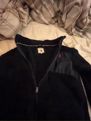 Svart fleecejacka från U.S. Polo Assn. - Mysig svart fleecejacka med hel dragkedja från U.S. Polo Assn. Bröstficka i nylon med röd logga broderad på vänster sida. Hög krage och enkel, stilren design som passar perfekt till lager-på-lager.