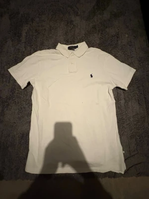 Vit pikétröja från Polo Ralph Lauren - Säljer en klassisk vit pikétröja från Polo Ralph Lauren med broderad logga på bröstet. Tröjan har krage, korta ärmar och knappar vid halsen. Perfekt för dig som gillar stilrena plagg och ikoniska märken.