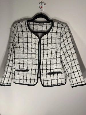 Vit rutig kavaj från Cellbes - Stilren vit kavaj från Cellbes med svart rutmönster och svarta kantdetaljer. Modellen är kraglös med två dekorativa fickor framtill och har en klassisk, rak passform. Materialet är strukturerat och ger en trendig look.