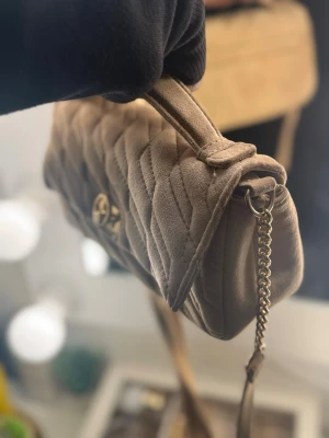 Beige axelväska - Säljer en beige quiltad handväska med guldfärgad kedja och vridlås. Väskan har en mjuk sammetsliknande yta och ett elegant mönster. Perfekt för dig som gillar stilrena accessoarer med en lyxig touch. 