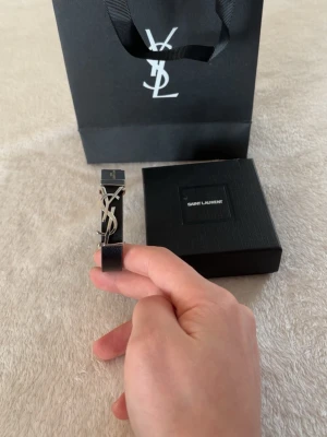 Saint Laurent svart läderarmband - Snyggt svart armband från Saint Laurent i äkta läder med silverfärgat YSL-logo och metallspänne där det står 'Saint Laurent Paris'. Stilren och exklusiv design som passar perfekt till din outfit.