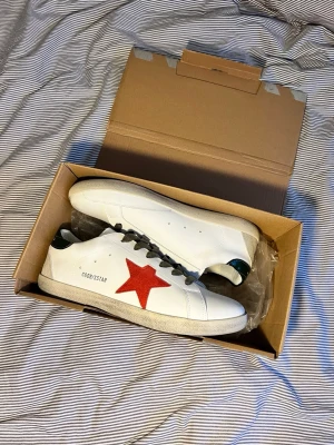 Golden Goose vita sneakers med röd stjärna - Snygga vita sneakers från Golden Goose med en ikonisk röd stjärna på sidan och svarta skosnören. Skorna har detaljer i grönt på hälen och GGDB/SSTAR-tryck på sidan. Tillverkade i läder med slitstark sula och klassisk låg profil. 0bs; lnte 0rglnal‼️, men mycket bra kvalitet 