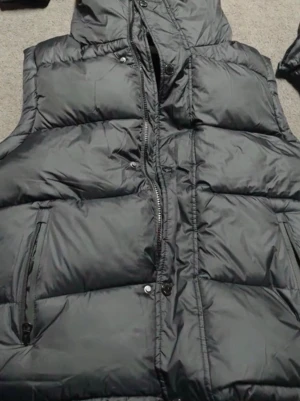 Svart dunväst med huva - Stilren svart dunväst med huva och dragkedja framtill. Västen har två fickor med dragkedja och ett rutigt innerfoder i svart, vitt och rött. Perfekt för lager-på-lager och håller dig varm under kyliga dagar.