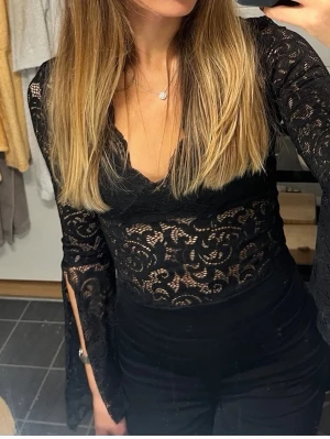 Svart spetsblus med v-ringning - Säljer en svart, långärmad spetsblus med v-ringning och transparent mönster. Blusen har figurnära passform och slitsade ärmar som ger en extra snygg detalj. Perfekt för dig som vill ha en trendig och elegant topp med coola spetsdetaljer.