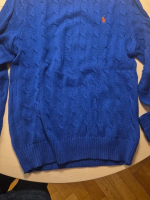 Blå kabelstickad tröja Polo Ralph Lauren - Säljer en blå kabelstickad tröja från Polo Ralph Lauren med orange broderad logga på bröstet. Använd 1 gång