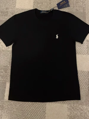 Svart t-shirt från Polo Ralph Lauren - Klassisk svart t-shirt från Polo Ralph Lauren med den ikoniska vita logon broderad på bröstet. T-shirten har rund hals och korta ärmar, tillverkad i mjuk bomull för en skön känsla. Perfekt till jeans eller shorts. Använd 1 gång bara, tagen ingår också vid frakt men den är inte satt på bara.