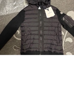 Svart pufferjacka från Moncler - Snygg svart pufferjacka från Moncler med stickade ärmar och huva. Jackan har två framfickor med tryckknappar, ribbade muddar och dragkedja framtill. Klassisk Moncler-logga på ärmen och quiltad kropp i glansigt material.