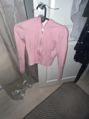 Rosa stickad hoodie med dragkedja - Säljer en söt rosa stickad hoodie med dragkedja framtill. Tröjan har långa ärmar, ribbstickad midja och är croppad i modellen. Perfekt för dig som gillar en avslappnad men trendig stil.