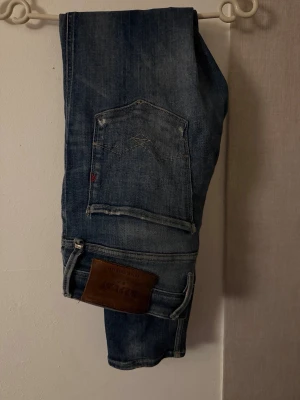 Replay anbass - Säljer ett par blå jeans från Replay med slitningar och distressed detaljer på framsidan. Jeansen har klassisk femficksdesign, läderpatch bak och är tillverkade i denim. Perfekt för dig som gillar en avslappnad och trendig look. Liten fläck där på baksidan som man ser på sista bilden. Även litet lite hål vid skrevet men inget man tänker på. Kan går ner i pris bara att höra av sig.   STORLEK. W31-L30