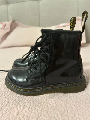 Svarta lackade stövletter Dr. Martens - Säljer ett par svarta lackade stövletter från Dr. Martens med klassisk gul söm, snörning framtill och dragflik bak. Skorna har en robust sula och den ikoniska Dr. Martens-looken. Perfekta för dig som vill ha en edgy och trendig stil.