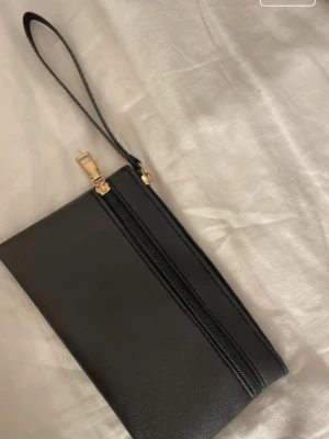 Svart plånbok med handledsrem - Stilren svart plånbok i slätt material med dragkedja och guldfärgad detalj. Praktisk handledsrem gör den enkel att bära med sig. Perfekt för att hålla koll på kort, cash och småprylar.