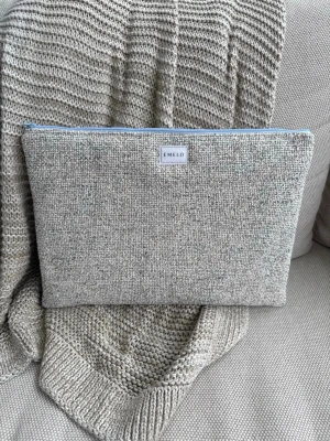 Beige stickad datorväska från Emelo - Snygg beige datorväska från Emelo i vävt tyg med mjuk känsla. Väskan har en enkel, rektangulär form och stängs med ett ljusblått blixtlås. Perfekt för att skydda din laptop med stil och en clean look. Passar laptop med maxmått: 33x24cm