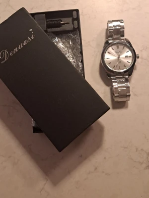 Silverfärgad klocka från Denuasi - !!HELT NY!!  Stilren klocka från Denuasi med silverfärgad länkarmband och rund urtavla. Urtavlan är också silverfärgad med tydliga markeringar, datumvisning och tre visare. Klassisk design som passar till många stilar. Levereras i originalask.