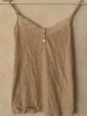 Beige linne med spets - Snygg beige linnetopp med smala axelband och spetskant upptill. Toppen har dekorativa knappar framtill och är tillverkad i mjukt bomullsmaterial. Perfekt att styla lager på lager eller bära som den är.