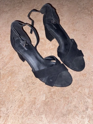 Svarta pumps med blockklack  - Svarta pumps med bred blockklack och öppen tå. Skorna har korsade remmar framtill och justerbar ankelrem för extra stöd. 