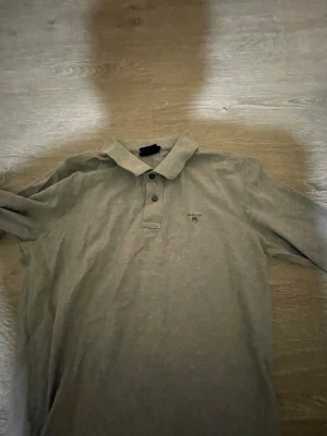 Grå långärmad skjorta från Gant - Klassisk grå långärmad skjorta från Gant med krage och knappar framtill. Skjortan har en diskret broderad logga på bröstet och är tillverkad i mjukt bomullsmaterial. Perfekt för en clean och stilren look.