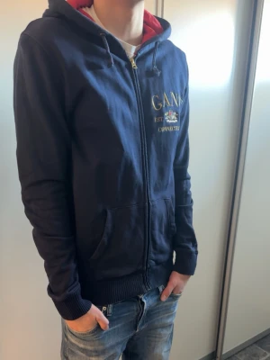 Marinblå hoodie från GANT - Snygg marinblå hoodie från GANT med dragkedja framtill och broderad logga på bröstet. Perfekt för dig som gillar vintage kläder.