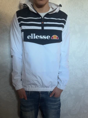 Vit vindjacka från Ellesse  - Snygg vit vindjacka från Ellesse med svart panel upptill och vita ränder. Perfekt för en sportig och avslappnad stil.