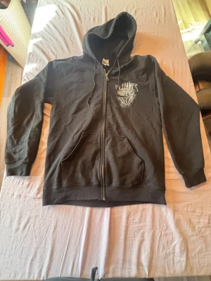 Svart In Flames zip hoodie - Säljer en svart zip hoodie från In Flames med tryck på både bröstet och ryggen. Hoodien har huva med dragsko, två fickor fram och ribbade muddar. Perfekt för dig som gillar bandmerch och vill ha en skön och stilren hoodie.