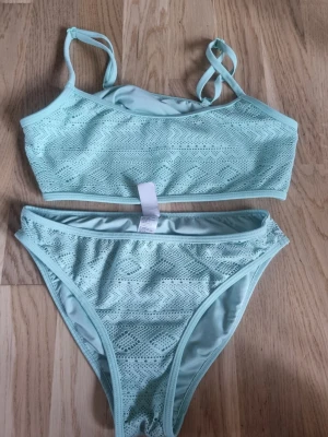 Turkos bikini med spetsmönster - Supersnygg turkos bikini med spetsliknande mönster. Toppen har smala axelband och underdelen har klassisk midja. Perfekt för strandhäng eller poolparty i sommar! Strl 158/164