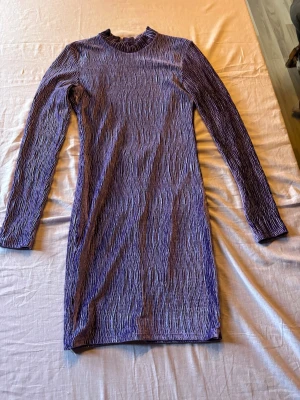 Lila långärmad kortklänning - Trendig lila kortklänning med lång ärm och cool struktur i tyget. Klänningen har en hög hals och en snygg öppen rygg som ger en extra twist. Perfekt för dig som vill sticka ut med en unik look.