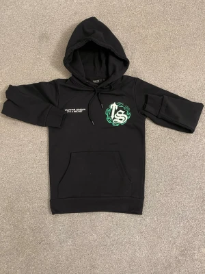Svart hoodie Trapstar London - Svart hoodie från Trapstar London med stor grön och vit grafisk logga på bröstet och en stor kedjedesign med bokstäver på ryggen. Klassisk känguruficka framtill och justerbar huva med snören. Tillverkad i mjukt material för en avslappnad look.