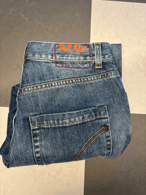Blå slitna jeans från dondup - Säljer ett par blå jeans från dondups med slitningar på låren och knäna. Jeansen har klassisk femficksmodell, orange dondup-logga bak och kontrastsömmar. Perfekta för en avslappnad och trendig look.