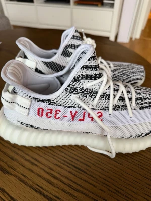 Yeezy Boost 350 Zebra sneakers - Säljer ett par Adidas Yeezy Boost 350 Zebra med svartvitt mönster och röda SPLY-350-detaljer på sidan. Skorna har en stickad ovandel, vita snören och en ribbad, vit sula i Boost-material. Perfekta för dig som gillar streetwear och vill sticka ut. Man får även med en gratis sak på sista bilden🔥🤯