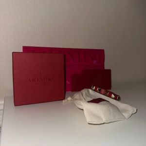Valentino rött läderarmband med nitar - Säljer ett snyggt rött armband från Valentino i äkta läder med guldiga pyramidformade nitar runt om. Armbandet har justerbart spänne och kommer med originalförpackning och dustbag. Perfekt accessoar för att lyfta din outfit. Finns en liten defekt vid spännet. ÄKTA. 