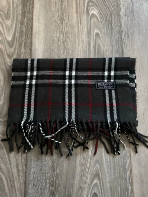 Rutig halsduk från Burberrys i ull - Klassisk rutig halsduk från Burberrys i 100% lammull. Färgerna är svart, vitt och rött i ett tidlöst rutmönster. Halsduken har fransar i ändarna och är tillverkad i England. Perfekt accessoar för att lyfta din outfit.