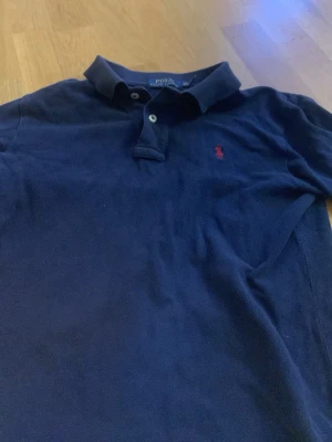 Marinblå piké från Polo Ralph Lauren - Klassisk marinblå pikétröja från Polo Ralph Lauren med röd broderad logga på bröstet. Modellen är Classic Fit och har krage samt knappar vid halsen. Perfekt för en stilren och avslappnad look.