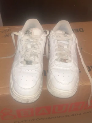 Vita Nike Air Force 1 sneakers - Klassiska vita Nike Air Force 1 sneakers med lågt skaft. Skorna har perforerad tå, vita skosnören och metallbricka med 'AF1' på snörningen. Tillverkade i läder med gummisula och diskret Nike-logga på sidan och tungan.