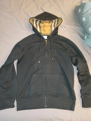 Svart hoodie med dragkedja Burberry - Snygg svart hoodie från Burberry med dragkedja och klassiskt rutigt foder i huvan. Har två stora fickor framtill och broderad logga på bröstet. 