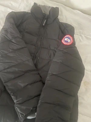 Svart pufferjacka från Canada Goose - Säljer en svart pufferjacka från Canada Goose med hög krage och dragkedja framtill. Jackan har det klassiska runda Canada Goose-märket på ärmen och är vadderad för extra värme. Perfekt för kalla vinterdagar.