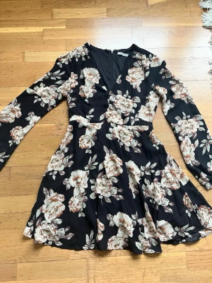 Blommig svart klänning Glamorous - Kort svart klänning från Glamorous med stora blommiga mönster i beige och ljusbrunt. Klänningen har v-ringning, långärmade ärmar och markerad midja. 