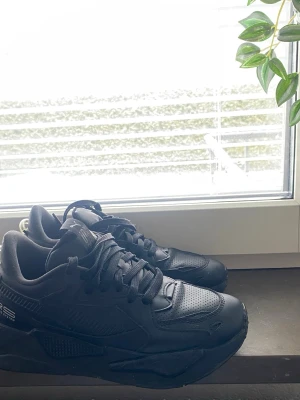 Svarta Puma RS-X sneakers - Säljer ett par helsvarta Puma RS-X sneakers med chunky sula och sportig design. Skorna har ovandel i syntet och mesh med RS-logga på sidan. Klassiska svarta skosnören och perforerade detaljer för extra stilpoäng.