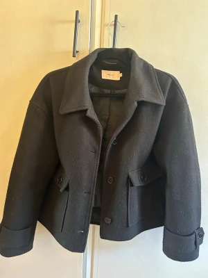 Kort svart ulljacka från Nelly - Stilren svart jacka från Nelly i ullblandning. Kort modell med bred krage, knäppning framtill och dekorativa fickor med knappar. Ärmslut med knappdetaljer ger en snygg touch. Perfekt för dig som gillar klassisk men trendig stil.