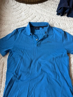 Blå pikétröja från GANT - Säljer en klassisk blå pikétröja från GANT med korta ärmar och krage. Tröjan har två knappar vid halsen och diskret GANT-logga broderad på bröstet. Tillverkad i mjuk bomull som känns skön mot huden. Man får även med en gratis vara som syns på sista bilden🤯🤯🤯