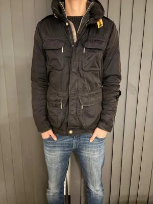 Svart Parajumper fieldjacka - Svart jacka från Parajumpers med flera fickor framtill, hög krage och gul detalj vid halsen. Jackan har en patch på ena ärmen och dragkedja samt knappar framtill. Perfekt för kyligare dagar.