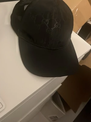 Svart Gucci keps med mesh - Säljer en svart keps från Gucci med diskret GG-mönster på framsidan och mesh-paneler på sidorna. Klassisk böjd skärm och justerbar baksida. Perfekt för dig som vill ha en stilren accessoar med exklusiv känsla.
