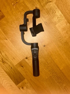 Svart gimbal för smartphone - Stabil gimbal i svart plast och metall för smartphone. Ergonomiskt handtag med kontrollpanel och joystick för enkel styrning. Perfekt för att filma stabila videos och ta snygga bilder med mobilen.