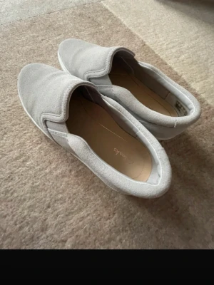 Ljusa loafers från Clarks i mocka (stl 37) - Säljer ett par ljusgrå loafers från Clarks i mjuk mocka. Skorna har en klassisk slip-on design med rundad tå och diskret söm längs kanten. Perfekta för dig som gillar stilrena och bekväma skor med enkel look.