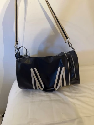 Fred Perry svart och vit resväska - Snygg svart och vit resväska från Fred Perry i cylinderformad modell. Ser nästan oanvänd ut och är smält använd Väskan har vita detaljer, bred axelrem med ränder och två handtag. Materialet är glansigt konstläder och loggan syns tydligt på sidan. Perfekt för weekendresor eller gymmet.