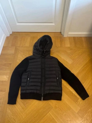 Svart Moncler cardigan - Säljer en svart cardigan från Moncler med stickade ärmar och huva. Jackan har två fickor med dragkedja framtill och en extra ficka på ärmen med Moncler-logga. Kroppen är quiltad och vadderad medan ärmarna är ribbstickade för extra komfort.