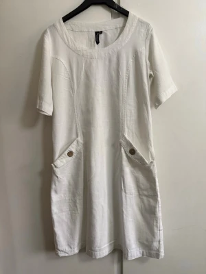 Vit kort klänning i linne från SIGNATURE - Vit kort klänning i linne från Signature med rund halsringning, korta ärmar och två stora fickor framtill med knappdetaljer. Enkel och stilren design som passar perfekt till sommaren.