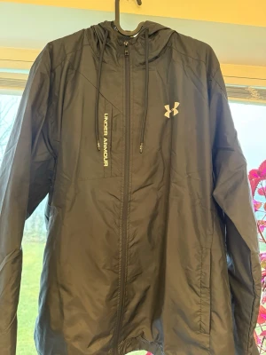 Svart vindjacka från Under Armour - Snygg svart vindjacka från Under Armour med dragkedja framtill, justerbar huva och logga på bröstet. Jackan är tillverkad i lätt och tåligt material, perfekt för blåsiga dagar. Sportig look med diskreta detaljer och praktiska fickor. Pris går att dissuktera. Vill man ha fler bilder så är det bara att säga till.