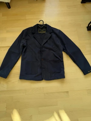 Mörkblå ullkavaj  - Passar L/M. Snygg mörkblå kavaj/overshirt i ullmaterial. Klassisk design med två framfickor, enkelknäppning och broderad detalj på ärmen. Perfekt för dig som vill ha en stilren och bekväm blazer med modern touch.