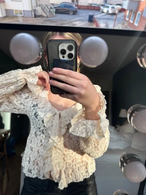 Vit spetsblus med volanger - Romantisk vit spetsblus med volanger längs knäppningen och vid ärmsluten. Blusen har v-ringning, pärlknappar framtill och är tillverkad i ett genombrutet spetsmaterial som ger en vintagekänsla. Perfekt för dig som gillar detaljer och en feminin stil. Prislapp kvar! Ord pris 349kr